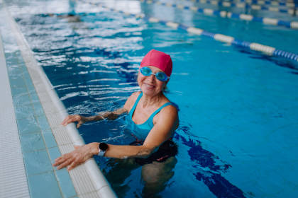 Happy senior woman in swimming pool, leaning on edge and looking at camera.