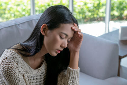 Headache. Young Asian woman is sitting on a sofa with her eyes closed.