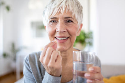 Smiling happy healthy middle aged 50s woman holding glass of water taking dietary supplement vitamin pill. Old women multivitamins antioxidants for anti age beauty.